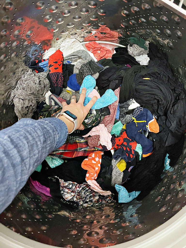 Author's left hand reaching into a washing machine to grab a handful of colorful clothing items